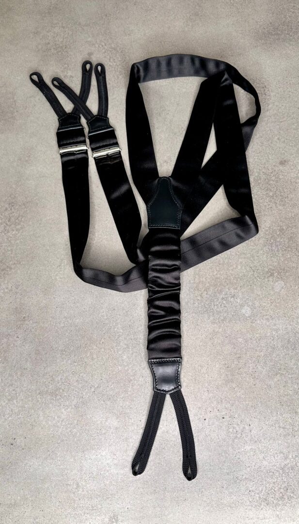 A black Barathea Silk Tuxedo Suspenders - Made in Italy with adjustable ends and metal buckles is laid out on a grey, textured surface, resembling the neat precision of a folded handkerchief.
