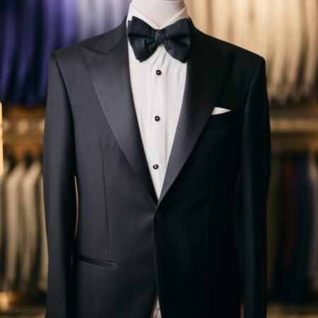 A Black Tuxedo - Made in Italy with a white dress shirt and black bow tie elegantly adorns a mannequin in a store, surrounded by other meticulously crafted suits hanging in the background.