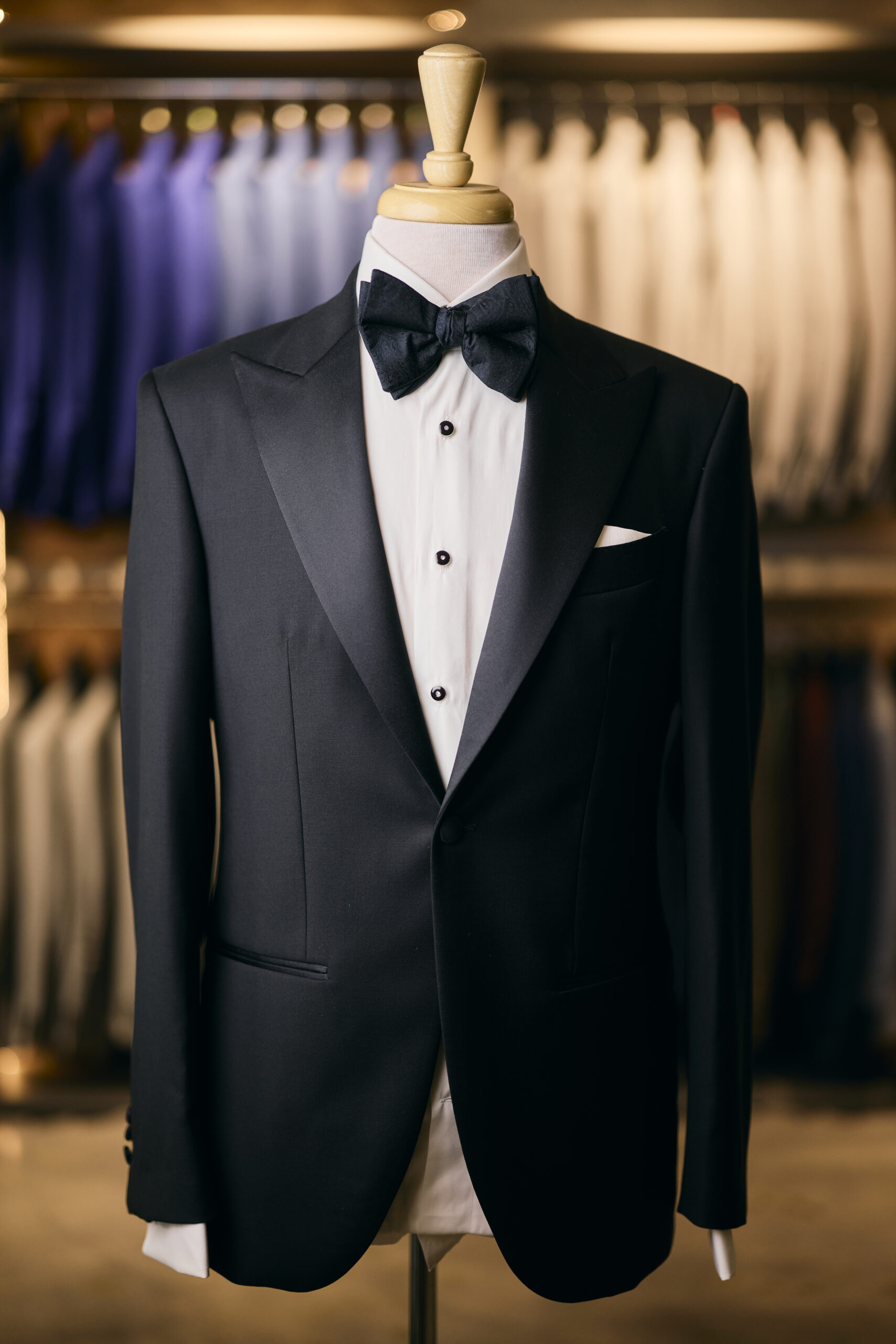 A Black Tuxedo - Made in Italy with a white dress shirt and black bow tie elegantly adorns a mannequin in a store, surrounded by other meticulously crafted suits hanging in the background.